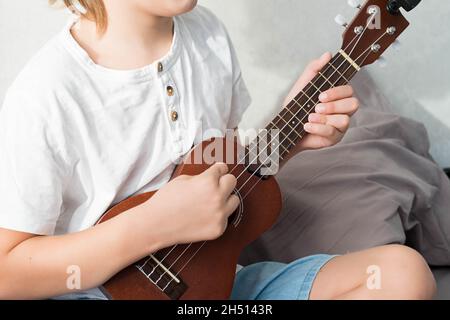 Ragazzo giovane che sintonizza l'ukulele a casa. Ragazzo con capelli biondi seduto sul divano suonando chitarra acustica. Foto Stock