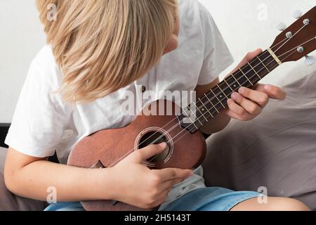 Ragazzo giovane che sintonizza l'ukulele a casa. Ragazzo con capelli biondi seduto sul divano suonando chitarra acustica. Foto Stock