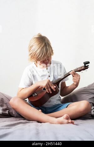 Ragazzo giovane che sintonizza l'ukulele a casa. Ragazzo con capelli biondi seduto sul divano suonando chitarra acustica. Foto Stock