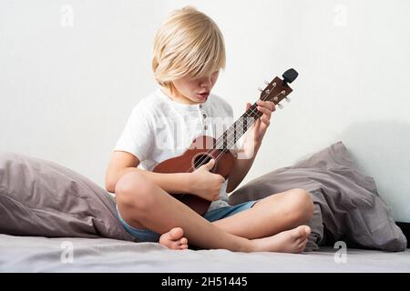 Ragazzo giovane che sintonizza l'ukulele a casa. Ragazzo con capelli biondi seduto sul divano suonando chitarra acustica. Foto Stock