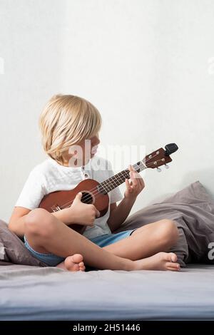 Ragazzo giovane che sintonizza l'ukulele a casa. Ragazzo con capelli biondi seduto sul divano suonando chitarra acustica. Foto Stock