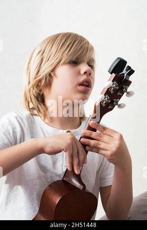 Ragazzo giovane che sintonizza l'ukulele a casa. Ragazzo con capelli biondi seduto sul divano suonando chitarra acustica. Foto Stock