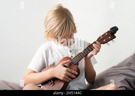 Ragazzo giovane che sintonizza l'ukulele a casa. Ragazzo con capelli biondi seduto sul divano suonando chitarra acustica. Foto Stock