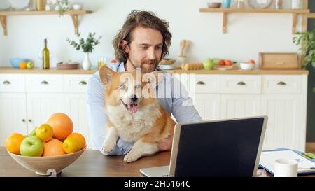 Uomo attraente che lavora con un notebook da casa. Giovane uomo con setole seduto al tavolo e tenendo in braccio un cane carino durante il lavoro a distanza Foto Stock