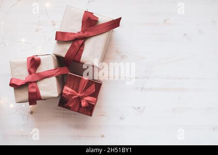 Tre regali di Natale con archi a nastro rossi e piccole luci bianche di Natale bordo su sfondo di legno bianco con spazio copia Foto Stock