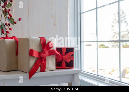 Regali di Natale con archi a nastro rosso su sfondo bianco seduti su panca da finestra in casa con bacche rosse e luci bianche con spazio copia Foto Stock