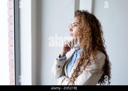 Donna d'affari biraciale pensata che guarda attraverso la finestra mentre si alza con la mano sul mento in ufficio Foto Stock