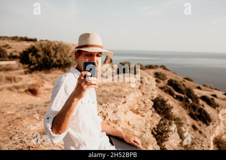 Digital nomad, uomo nel cappello, un uomo d'affari con un laptop siede sulle rocce vicino al mare durante il tramonto, effettua una transazione di affari in linea da a. Foto Stock