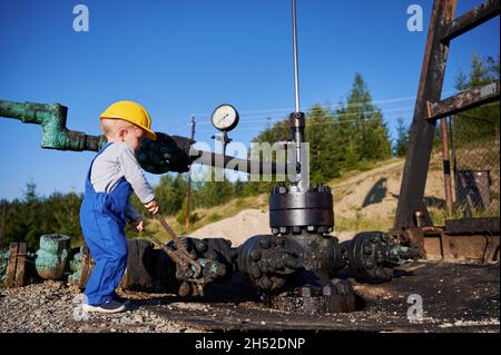 Attrezzo per l'esame del preschooler maschio per dadi e bulloni vicino alla tubazione dell'olio. Ragazzino in abiti da lavoro speciali che imparano meccanica, provando a professione di oilman. Foto Stock