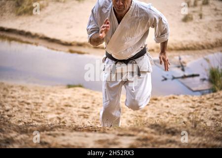 Karate uomo con cintura nera in un vecchio kimono corre su una collina di sabbia. Un piccolo fiume scorre sotto. Concetto. Foto Stock
