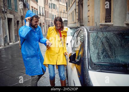 Una giovane coppia allegra in impermeabile ha raggiunto l'auto mentre si è allontanata dalla pioggia in una giornata nuvolosa in città. Passeggiata, pioggia, città, rapporto Foto Stock