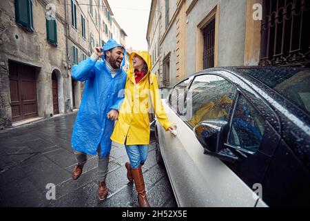 Una giovane coppia innamorata è entusiasta mentre corre in macchina mentre si fa una passeggiata in città in impermeabile in un'atmosfera allegra durante una giornata piovosa. Passeggiata, Foto Stock