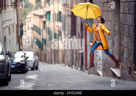 Una giovane ragazza con un impermeabile giallo e un ombrello salta sulla strada mentre cammina per la città in modo allegro in una giornata piovosa. Passeggiata, pioggia, città Foto Stock