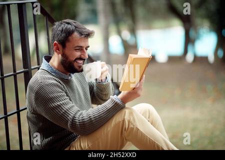 Un giovane e bello uomo sta gustando un libro e un caffè mentre si siede sulle scale di fronte alla casa in un'atmosfera rilassata. Città, fuori, riposo Foto Stock