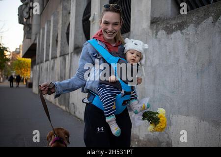 Belgrado, 31 ottobre 2021: Una giovane donna con un bambino in un portatore e un ampio sorriso cammina cane lungo la strada portando un bouquet di fiori gialli Foto Stock