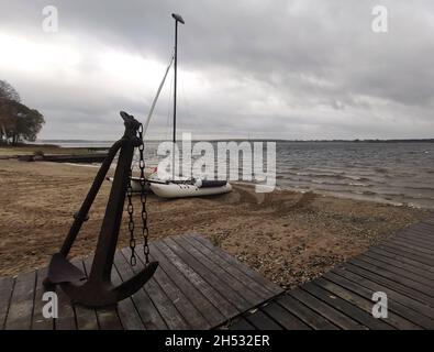 Grande vecchia arrugginita ancora su molo di legno sulla riva sabbiosa del fiume Volga con barca gonfiabile e il fiume stesso sullo sfondo Foto Stock