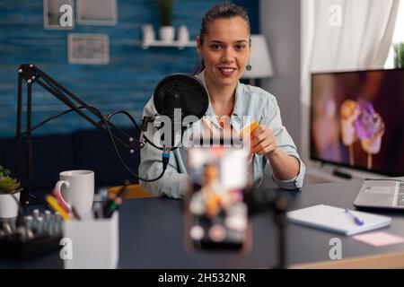 Influencer registra la recensione di un libro giallo per il canale dei social media. Autore di contenuti donna che presenta consigli sui prodotti per il vlog online in casa studio con microfono e laptop. Foto Stock