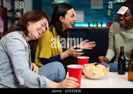 Ridendo amici multietnici che giocano a chi sto giocando utilizzando le note stickey godendo di passare il tempo insieme durante la festa wekeend a casa. Gruppo di persone che bevono birra indovinando la risposta corretta Foto Stock