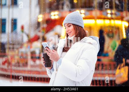 La vista laterale della donna felice indossa in giacche legge smartphone alla città di Natale, mentre tiene il caffè Foto Stock