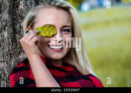 la ragazza bionda appoggiata la testa contro un albero, nasconde giocosamente un occhio dietro una foglia, amando la natura Foto Stock