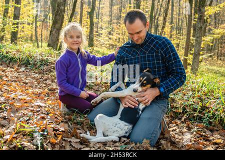 Un uomo in plaid caffè e jeans con un cane in grembo si siede accanto a una ragazza. Il bambino guarda nella fotocamera e sorride. Padre e figlia in un autum Foto Stock