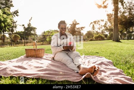 Donna incinta africana che usa lo smartphone mobile mentre fa un picnic nel parco - concetto di stile di vita di maternità Foto Stock