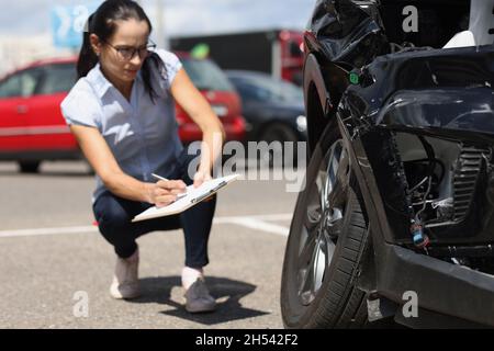 Ispettore donna che scrive in documenti su appunti vicino auto rotta. Assicurazione auto Foto Stock