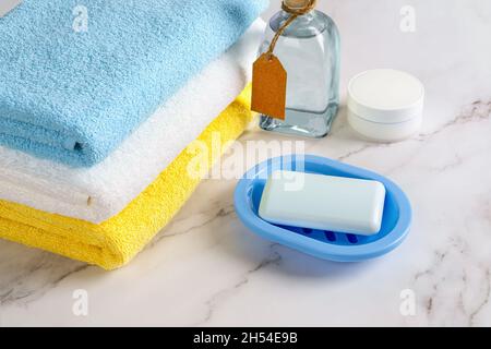 Sapone su un piatto blu, crema idratante per la pelle e lozione tonica vicino a una pila di asciugamani in spugna di cotone sopra la scrivania di marmo. Concetti di igiene, purezza e prodotti da bagno. Foto Stock