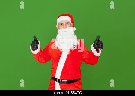 Un uomo caucasico vestito come Babbo Natale facendo un gesto alzando i pollici delle sue due mani. Lo sfondo è verde. Foto Stock