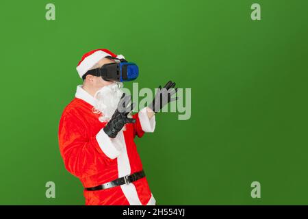 Un uomo caucasico vestito come Babbo Natale indossa occhiali di realtà virtuale e si diverta a giocare con loro. Lo sfondo è verde. Foto Stock