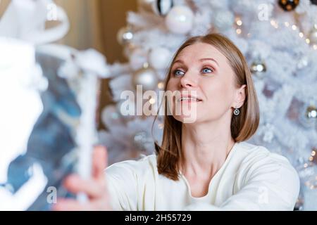 Ritratto di donna caucasica sorridente che tiene la scatola regalo d'argento vicino all'albero. Una giovane donna apre il suo presente. Bella ragazza felice sotto l'albero di natale. Foto Stock