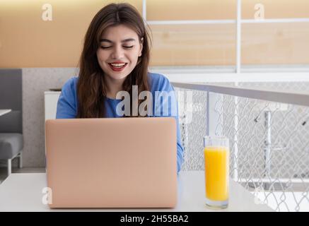Bella giovane donna in un maglione blu lavora a un computer portatile in un bar. Foto Stock