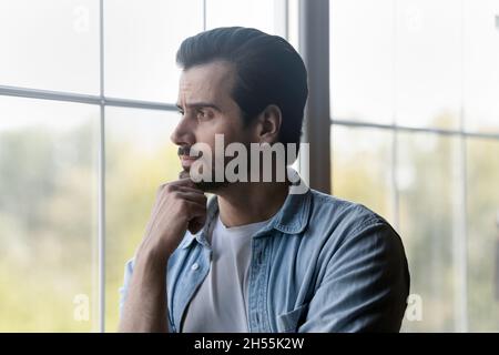 Il tipo di bearded del millennial pensivo sta dalla finestra osserva la distanza Foto Stock