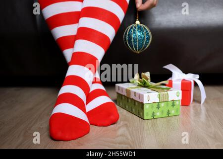 Gambe femminili in calze di Natale ginocchio su un pavimento vicino alle scatole regalo. Donna seduta su un letto con palla giocattolo in mano, festa di Capodanno Foto Stock