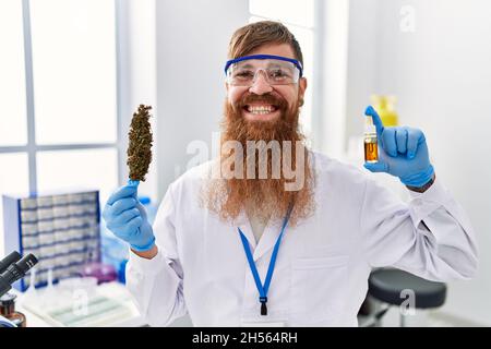 Uomo rosso con barba lunga che lavora presso il laboratorio di scienziato tenendo erbacce e olio cbd sorridendo con un sorriso felice e fresco sul viso. Mostrando denti. Foto Stock