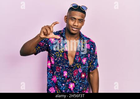 Giovane uomo nero che indossa una camicia hawaiana e occhiali da sole sorridenti e fiduciosi gesturing con mano facendo piccolo segno di taglia con le dita guardando e la camma Foto Stock