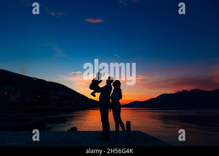 Silhouette di padre che tiene piccolo bambino e madre in piedi accanto a loro sul molo, sullo sfondo del mare, montagne e case al tramonto Foto Stock