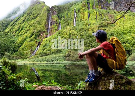 Ragazzo seduto su un tronco d'albero al lago Poo Ribeira do Ferreiro, guardando le cascate, Poco da Alagoinha, Fajãzinha, Flores Island, Azzorre Foto Stock
