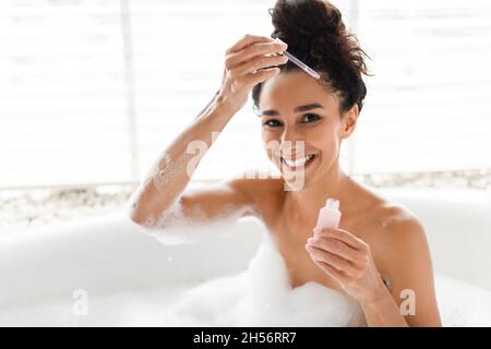 Giovane donna che applica siero idratante sul viso mentre si rilassa in bagno schiumoso, facendo il trattamento di bellezza a casa Foto Stock