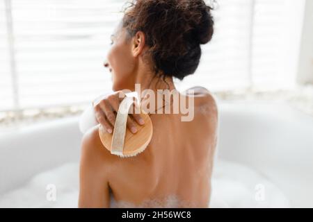 Vista posteriore della giovane donna che fa massaggio linfatico con spazzola, strofinando la sua pelle in bagno schiumoso a casa Foto Stock