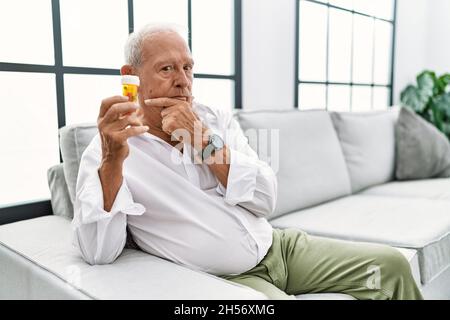 Uomo anziano che tiene pillole guardando fiducioso la macchina fotografica sorridendo con le braccia incrociate e la mano sollevata sul mento. Pensare positivo. Foto Stock