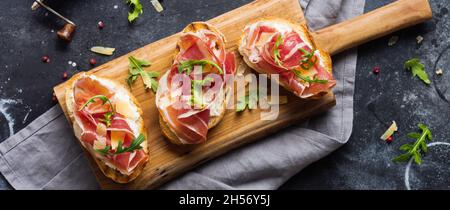 Sandwich al prosciutto aperto, rucola e formaggio duro, serviti su stand di legno con aglass di vino rosso su sfondo scuro vecchio aconcrete. Stile Rustik. Vista dall'alto. Foto Stock