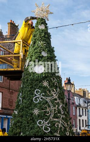 Dunbar, East Lothian, Scozia, Regno Unito, 7 novembre 2021. Luci di Natale erette: La High Street è chiusa per consentire alle luci di Natale di essere riposte attraverso la strada principale attraverso la città. Nella foto: Un uomo mette una stella sulla cima di un albero di Natale fuori dal municipio accanto alla statua di John Muir come un ragazzo Foto Stock