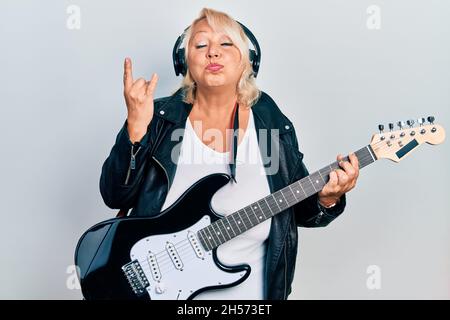 Donna bionda di mezza età che suona la chitarra elettrica facendo le corna segno con le dita puffing guance con faccia divertente. Bocca gonfiata con aria, cattura aria. Foto Stock