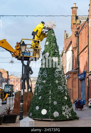 Dunbar, East Lothian, Scozia, Regno Unito, 7 novembre 2021. Luci di Natale erette: La High Street è chiusa per consentire alle luci di Natale di essere riposte attraverso la strada principale attraverso la città. Nella foto: Un uomo mette una stella sulla cima di un albero di Natale fuori dal municipio accanto alla statua di John Muir come un ragazzo Foto Stock