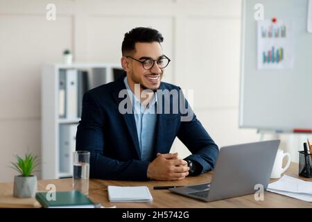 Sicuro giovane imprenditore arabo in abito formale utilizzando il computer portatile alla sua scrivania, avendo incontri virtuali in ufficio Foto Stock