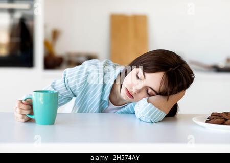 Giovane donna che dorme sul tavolo e tiene una tazza di caffè, si sente stanca, soffre di insonnia, seduta in cucina Foto Stock