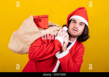 Un giovane vestito come Babbo Natale porta un sacco pieno di regali. Ha capelli lunghi, una barba e un'espressione di sorpresa. Foto Stock