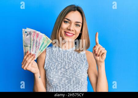 Giovane donna bionda caucasica in possesso di banconote dirham emirati arabi uniti sorridendo con un'idea o domanda puntando dito con volto felice, numero su Foto Stock