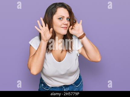 Giovane donna di più misura che indossa una t-shirt bianca casual cercando di sentire entrambe le mani sul gesto dell'orecchio, curioso per pettegolezzi. Problemi di udito, sordi Foto Stock